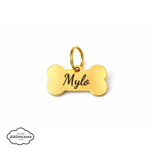 Könnte beinhalten: Ein goldfarbener, knochenförmiger Haustieranhänger mit dem Namen "Mylo" in schwarzer Schrift. Der Anhänger hat einen kleinen goldenen Ring zur Befestigung. Ein Haustierzubehör.