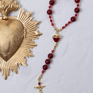 May include: A gold-colored religious heart-shaped pendant with a hinged door and a sunburst design. A rosary with red beads and a gold crucifix is also present. The rosary features a heart-shaped pendant and a gold crucifix.