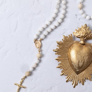 Puede incluir: Un rosario de cuentas blancas con detalles dorados y una cruz dorada. Tambi&eacute;n hay un colgante dorado del Sagrado Coraz&oacute;n con rayos de sol. Las cuentas del rosario son blancas y beige, y la cruz tiene un crucifijo.