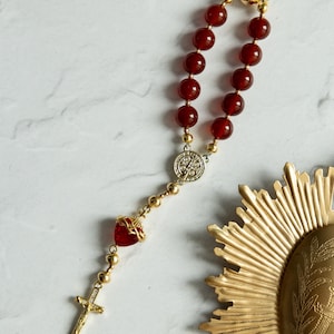 May include: A rosary featuring deep red beads and gold-coloured accents. The cross and connecting pieces are gold-toned, with a heart-shaped centerpiece. The rosary is set against a white marble background.