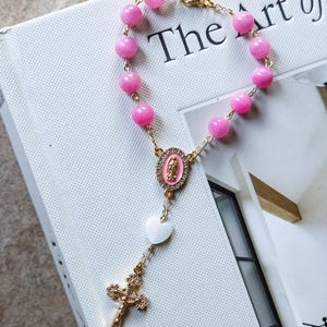 May include: A pink beaded rosary bracelet with gold-colored chain and accents. The bracelet features a gold cross, a white heart, and a pink oval pendant with a religious image. The beads are a vibrant pink color.