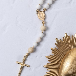 Puede incluir: Un rosario de cuentas blancas con detalles dorados, incluyendo un crucifijo y un medall&oacute;n central. El rosario presenta una mezcla de cuentas blancas y beige. Tambi&eacute;n hay un elemento decorativo dorado en forma de sol.