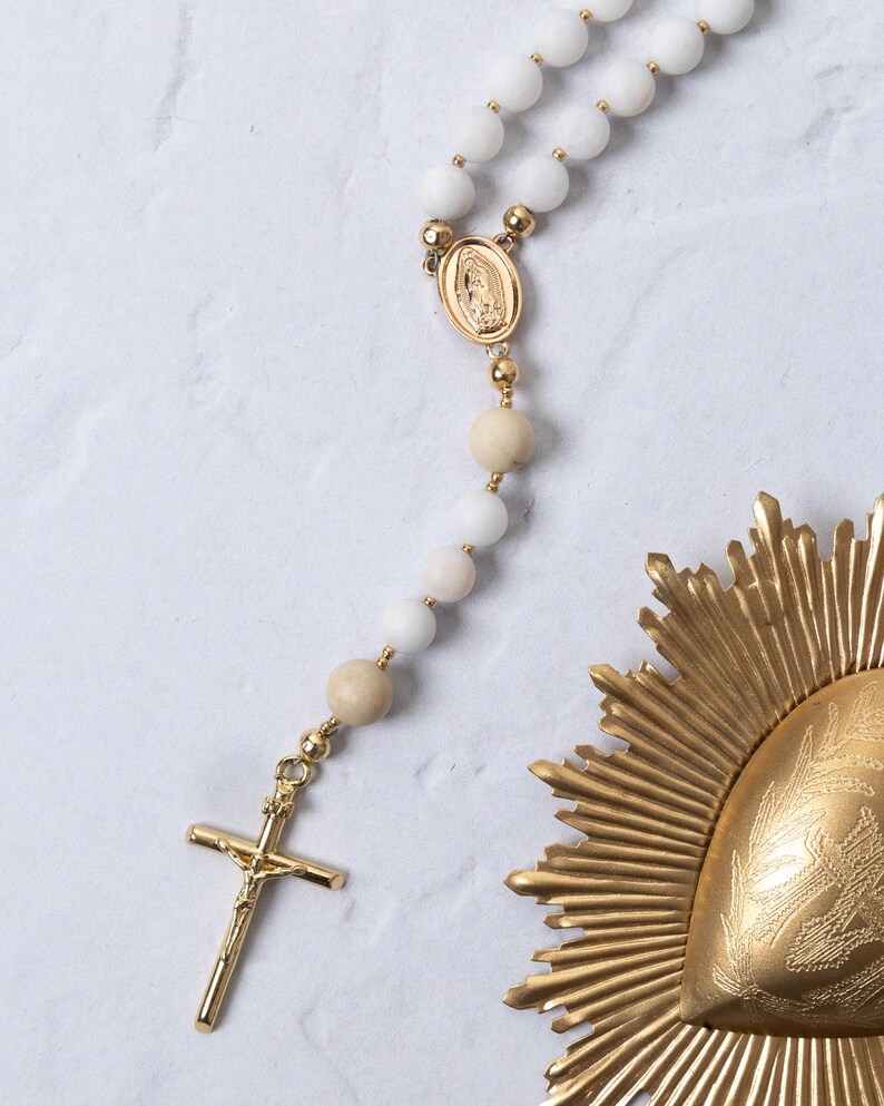 Puede incluir: Un rosario de cuentas blancas con detalles dorados, incluyendo un crucifijo y un medall&oacute;n central. El rosario presenta una mezcla de cuentas blancas y beige. Tambi&eacute;n hay un elemento decorativo dorado en forma de sol.