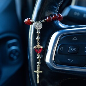 May include: A rosary featuring red beads, a silver centerpiece, a heart-shaped pendant, and a gold cross. The rosary is suspended from a black steering wheel. A religious item.