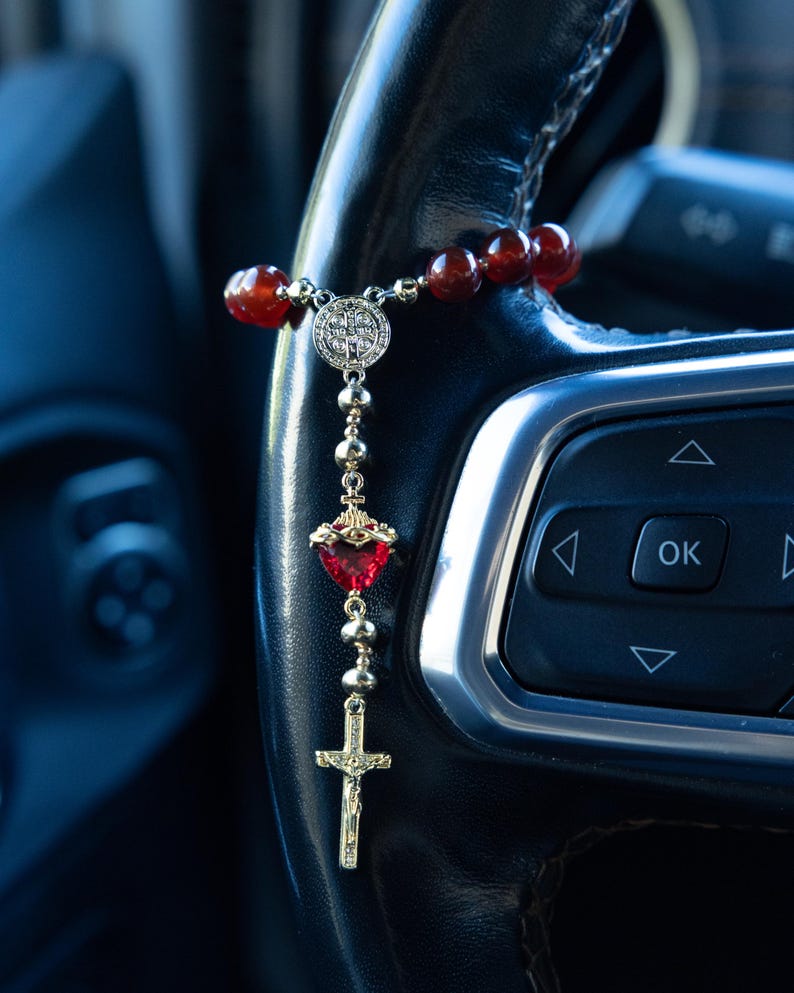 May include: A rosary featuring red beads, a silver centerpiece, a heart-shaped pendant, and a gold cross. The rosary is suspended from a black steering wheel. A religious item.
