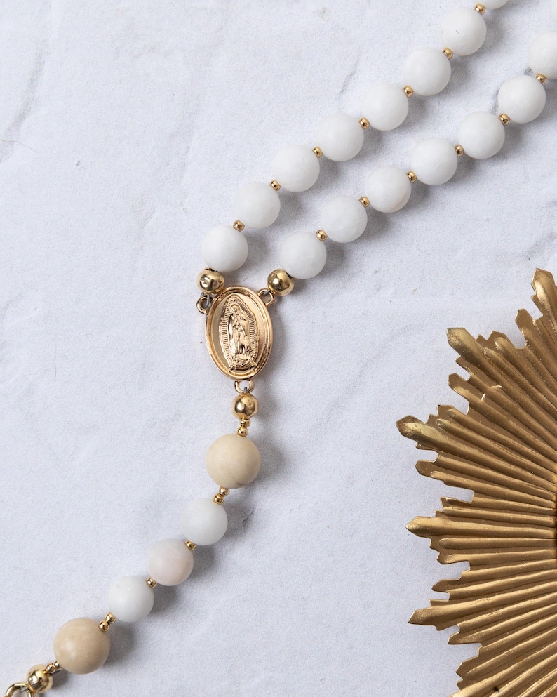 Puede incluir: Un rosario de cuentas blancas con detalles dorados y una pieza central dorada con una figura religiosa. Las cuentas son una mezcla de blanco y beige, con conectores dorados.