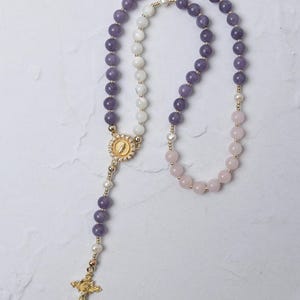 May include: A rosary featuring faceted purple beads, smooth white beads, and pale pink beads strung on gold-toned wire. The piece includes a central gold-toned medal with a circular relief design and a gold-toned crucifix at the bottom end.