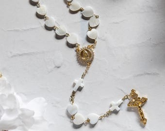 Puede incluir: Un rosario dorado con cuentas en forma de corazón y un crucifijo dorado. El rosario presenta una pieza central dorada con una figura religiosa y cuentas blancas en forma de cruz. Las cuentas son blancas, sobre un fondo de mármol.