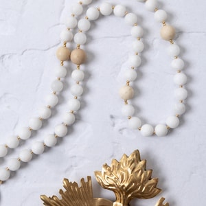 Puede incluir: Un collar de cuentas blancas con detalles dorados y cuentas de color beige. Tambi&eacute;n hay un colgante en forma de coraz&oacute;n de metal dorado con un dise&ntilde;o de llama. El collar se exhibe sobre una superficie de m&aacute;rmol blanco.