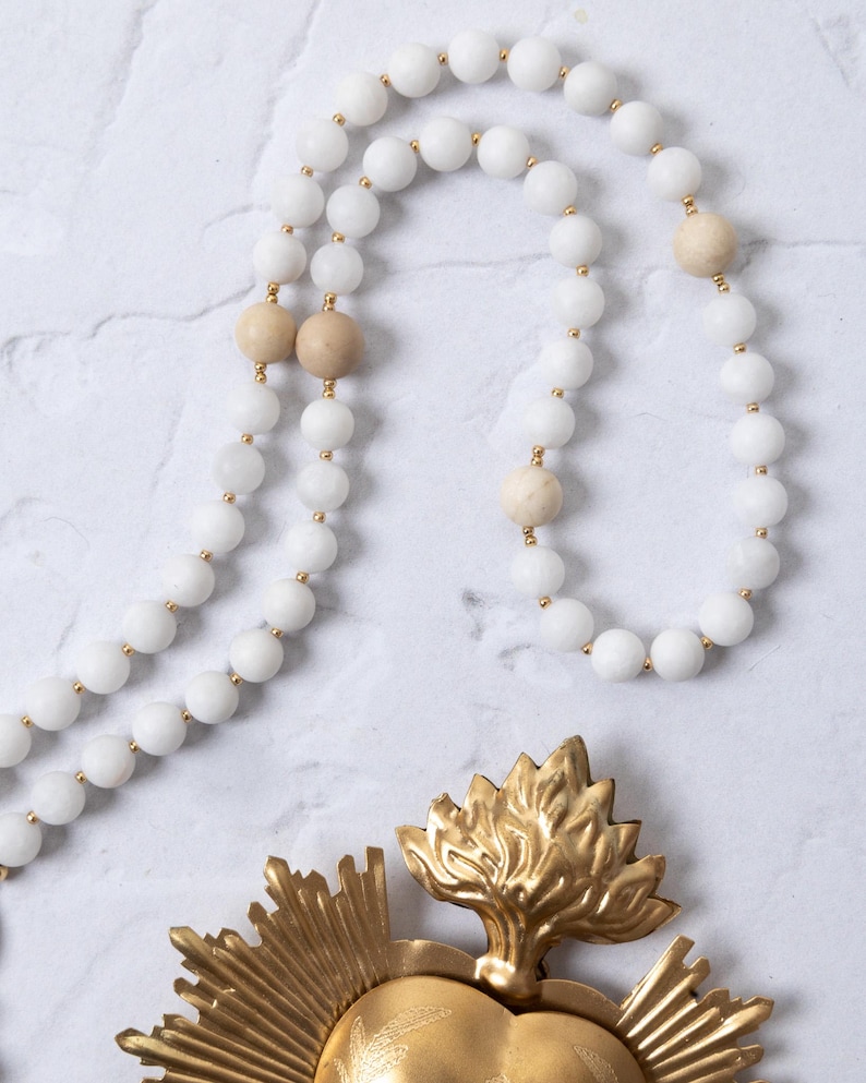 Puede incluir: Un collar de cuentas blancas con detalles dorados y cuentas de color beige. Tambi&eacute;n hay un colgante en forma de coraz&oacute;n de metal dorado con un dise&ntilde;o de llama. El collar se exhibe sobre una superficie de m&aacute;rmol blanco.