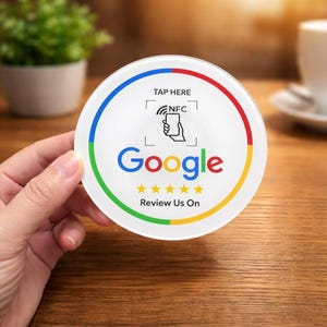 Może przedstawiać: Okrągła biała podkładka z logo Google w kolorach czerwonym, niebieskim, żółtym i zielonym. Podkładka zawiera tekst "TAP HERE" i "Review Us On". Zawiera również grafikę dłoni trzymającej telefon z symbolem NFC.