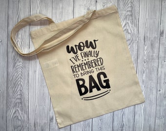 May include: A beige canvas tote bag with the text "Wow I've finally remembered to bring this bag" in black. The bag has two handles and is lying on a light gray wooden surface.