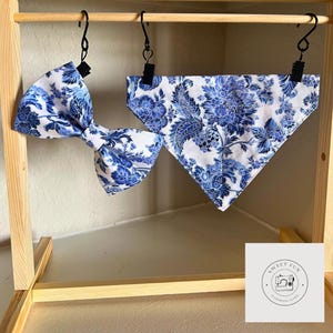 May include: A blue and white floral patterned bow tie and bandana set. The bandana is a triangle shape and the bow tie is a classic style. The set is hanging on a wooden rack with the text "Sweet Fun Handmade Items".
