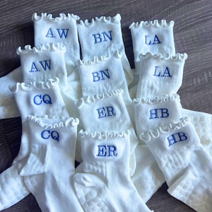 Könnte beinhalten: Eine Sammlung weißer Socken mit Rüschenbündchen, jede mit blauen Initialen bestickt. Die Socken haben ein strukturiertes Muster auf den Sohlen. Die Initialen sind AW, BN, LA, CQ, ER und HB.