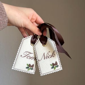 May include: Two cream-coloured gift tags with brown embroidered text reading "Tom" and "Nick". Each tag has a brown stitched border, a small pinecone design, and a brown ribbon. The tags are held by a person.