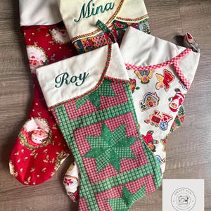 Op de afbeelding: Vier kerstsokken met witte manchetten en gepersonaliseerde namen geborduurd in groen. Eén sok heeft een kerstmanontwerp, een andere heeft een groen en rood sterrenpatroon en de andere hebben feestelijke patronen. De sokken liggen op een houten oppervlak.