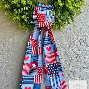 May include: A green wreath with a patriotic bow featuring red, white, and blue patchwork fabric. The bow has stars, stripes, and heart patterns. The wreath is made of green, leafy material.