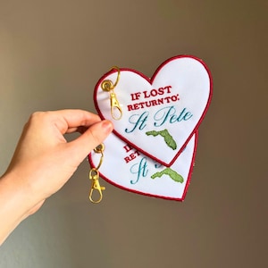 Puede incluir: Llavero bordado blanco en forma de corazón con ribete rojo y herrajes dorados. El llavero dice "IF LOST RETURN TO: St Pete" con un contorno bordado verde de Florida. El llavero está sostenido por una mano.