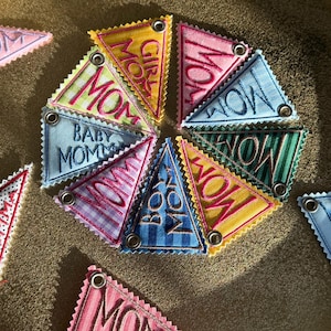 May include: A collection of colorful, triangular fabric patches with embroidered text. The patches feature phrases like "MOM," "GIRL MOM," and "BABY MOM." Each patch has a decorative zigzag edge and a metal grommet for hanging.