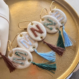 Puede incluir: Una colección de llaveros personalizados con etiquetas ovaladas de cuero blanquecino. Cada etiqueta presenta una letra o diseño en varios colores, con un cierre de metal dorado y una borla en tonos marrón, verde y azul. Los llaveros están colocados sobre una bandeja dorada.