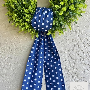 Op de afbeelding: Een groene buxuskrans met een marineblauwe strik met witte sterren. De strik is van stof en hangt aan de krans. De krans hangt aan een getextureerde muur.