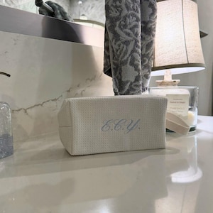 Puede incluir: Un neceser rectangular blanco roto con las iniciales "E.C.Y." bordadas en azul claro. La bolsa está hecha de una tela texturizada y se encuentra sobre una encimera blanca. Una toalla gris estampada cuelga cerca.