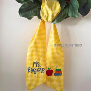 Könnte beinhalten: Ein gelb-weißes Gingham-Stoffband, das an einem Kranz aus grünen Blättern befestigt ist. Das Band zeigt den blauen Sticktext Ms. Rogers, einen roten Apfel, einen gelben Stift und drei gestapelte Bücher in Grün, Rot und Blau. Text @SWEETFUNSTUDIO.