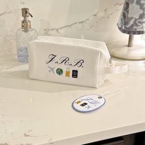 Puede incluir: Bolsa de viaje blanca con las iniciales "J.R.B." bordadas e iconos de viaje. Una etiqueta de equipaje a juego está al lado de la bolsa. La bolsa tiene un asa y un cierre de cremallera. Un dispensador de jabón transparente y una toalla están en el fondo.