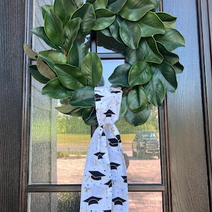 May include: A large, round wreath made of glossy, dark green magnolia leaves. A white ribbon with black graduation caps, gold stars, and yellow tassels hangs from the wreath. The wreath is hanging on a dark brown door with glass panels.