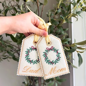 May include: Two cream-colored gift tags with embroidered Christmas wreath designs and the words "Dad" and "Mom" in gold script. Each tag has a gold ribbon for hanging.