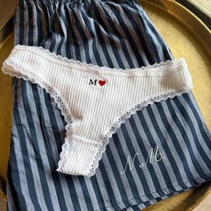 Puede incluir: Ropa interior blanca acanalada con ribete de encaje, bordada con la letra "M" y un corazón rojo. Descansa sobre un pantalón corto de pijama a rayas azules y blancas, bordado con las iniciales "N.M."