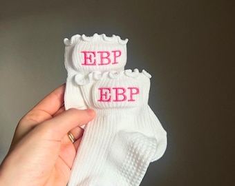 以下が含まれることがあります： 白いフリルのトップエッジが付いたアンクルソックスのペア。靴下にはピンク色の刺繍文字「EBP」があります。靴下は手で持たれています。