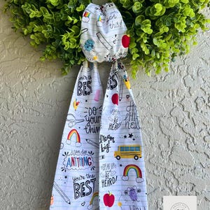 Könnte beinhalten: Ein grüner Kranz mit einer weißen Stoffschleife mit Illustrationen und Texten zum Thema Schule. Der Stoff enthält Bleistifte, Äpfel, Regenbogen und Sätze wie "You're the BEST" und "Speak Up Be Heard!"