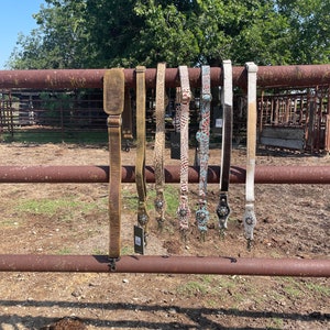 May include: A collection of leather and patterned straps, likely for a bag or guitar, hanging on a rusty metal fence. The straps vary in color, including brown, tan, and white, with decorative metal accents and buckles.