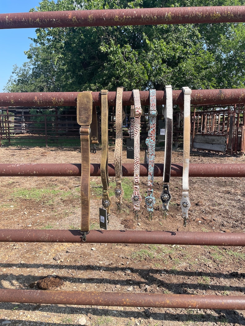 May include: A collection of leather and patterned straps, likely for a bag or guitar, hanging on a rusty metal fence. The straps vary in color, including brown, tan, and white, with decorative metal accents and buckles.