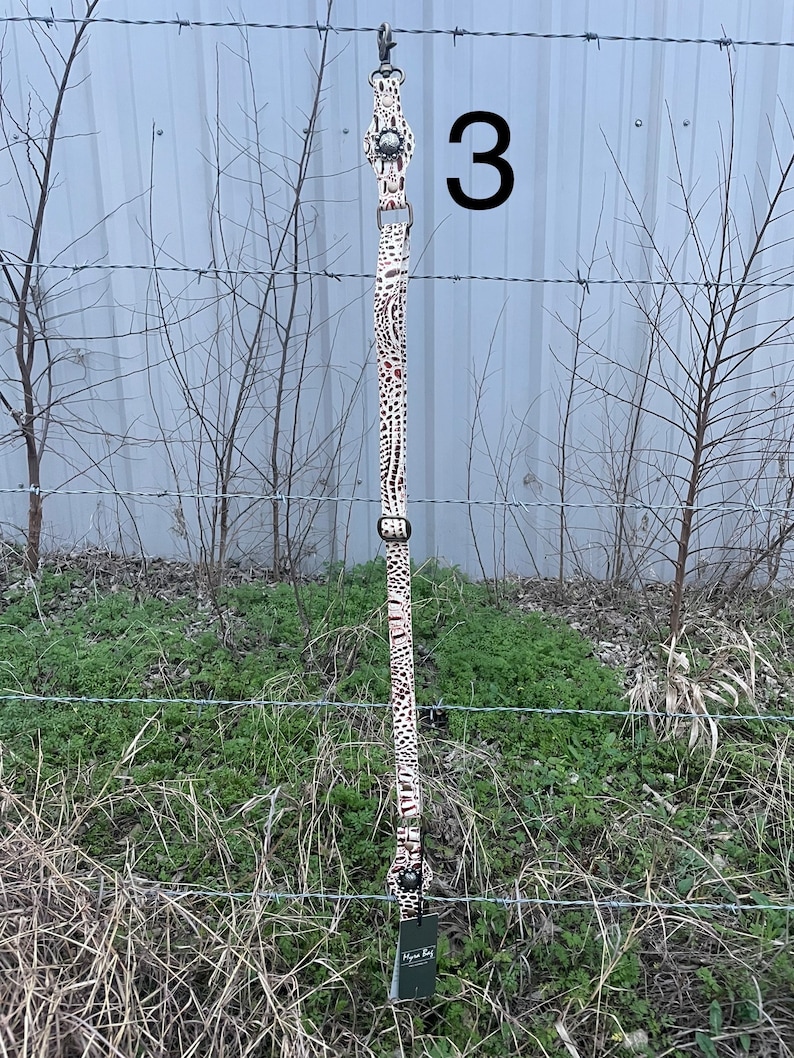 May include: A long, patterned strap with a metal clasp and hardware. The strap features a cream-colored background with brown and black spots. The number "3" is visible in the background. A tag with the text "Hyena Bay" is attached.
