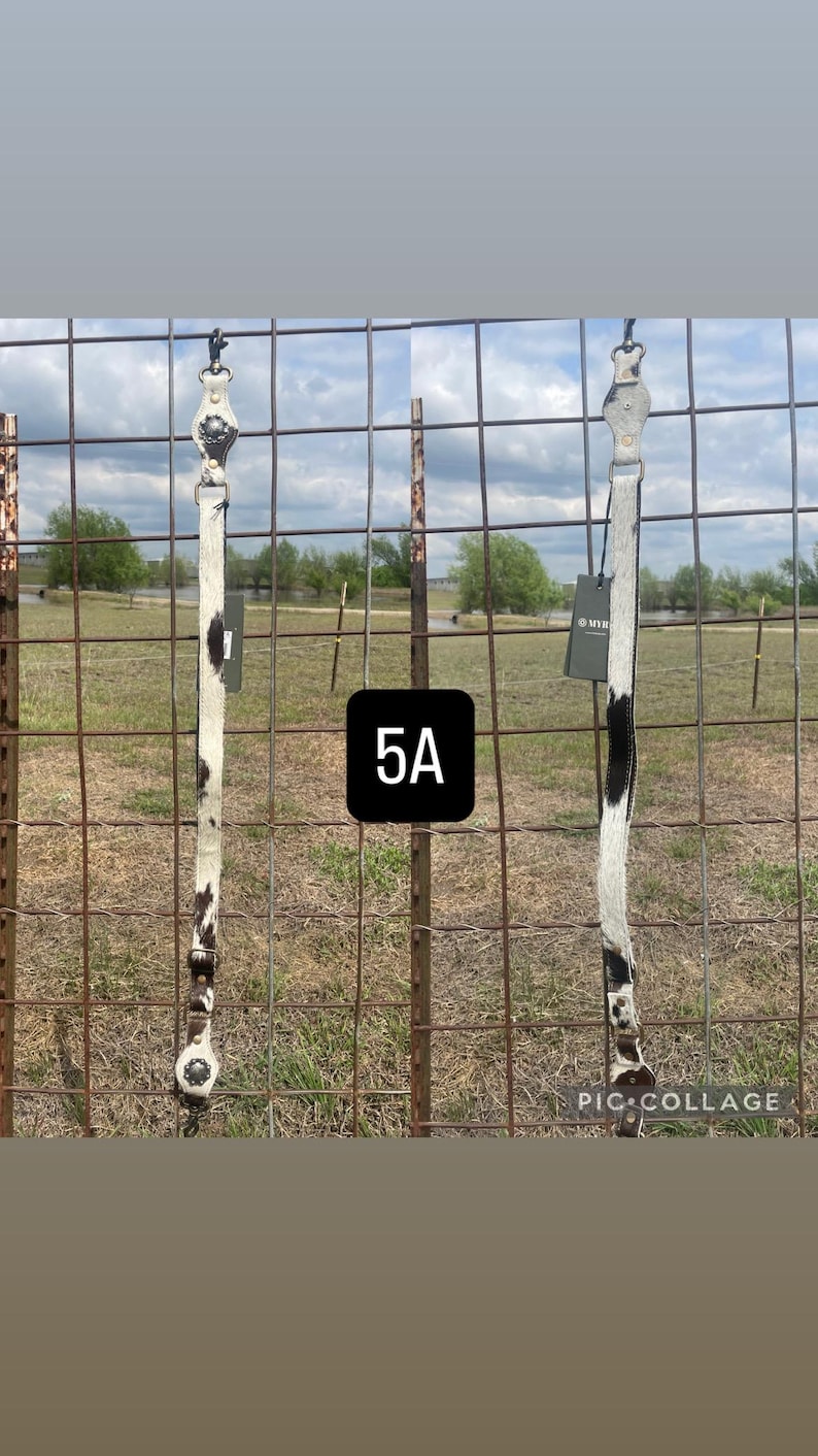 May include: Two leather horse tack pieces with a white and black cowhide pattern. The tack is hanging on a metal fence in front of a grassy field. The tag reads "MYR".