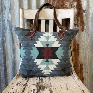 May include: A blue and white geometric patterned tote bag with brown leather handles. The bag is sitting on a white wooden chair.