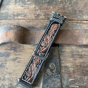 May include: Black leather watch band with a detailed floral design in brown and tan. The band features white stitching along the edges and a silver-tone buckle. The watch band is shown on a rustic wooden surface.