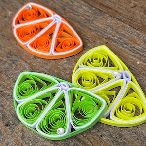 Puede incluir: Tres rodajas de cítricos de papel enrollado, naranja, limón y lima, con centros blancos y un adorno de strass en cada una.