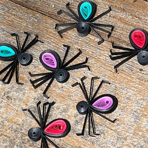 Op de afbeelding: Zes zwarte papieren spinnen met gekrulde papieren accenten in roze, paars en turquoise. De spinnen zijn gerangschikt op een houten oppervlak.