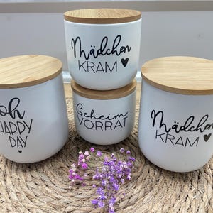 Könnte beinhalten: Drei weiße Keramikgläser mit Holzdeckeln. Die Gläser haben schwarze Beschriftungen mit den Worten "Oh Happy Day", "Mädchen Kram" und "Scheim Vorrat". Die Gläser stehen auf einer geflochtenen Strohmatte.