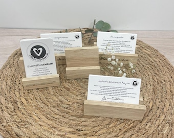 Könnte beinhalten: Mehrere Kartenhalter aus Holz, die weiße Karten mit Text und Logos zeigen. Die Karten enthalten Informationen und ein Herzlogo. Die Halter sind aus hellem Holz gefertigt und auf einer gewebten Matte angeordnet.