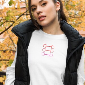 May include: A white long-sleeve shirt with a red, pink, and white design of four equestrian snaffle bits. The shirt is worn by a person with a black puffer vest.