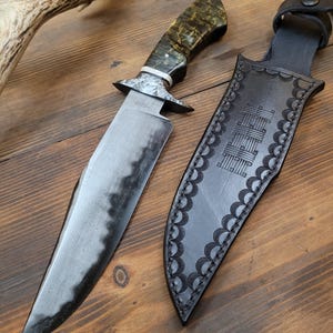 May include: A handcrafted hunting knife with a dark, textured blade and a patterned handle. The knife is displayed next to its black leather sheath, which has a decorative border and a belt loop. The knife and sheath are on a wooden surface.