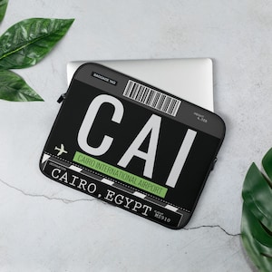 May include: Black laptop sleeve with a white luggage tag design. The tag reads "CAI CAIRO INTERNATIONAL AIRPORT CAIRO, EGYPT" with a barcode and flight number MS910.