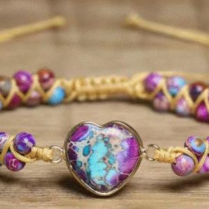 May include: A handmade bracelet featuring a heart-shaped pendant with a turquoise and purple pattern. The bracelet is made with colorful beads in shades of purple, blue, and pink, woven together with a tan cord. A unique piece of jewelry.