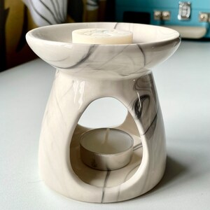 May include: White ceramic wax melt burner with a marble design. The burner has a round top with a small opening in the centre for a tea light. The burner is sitting on a white surface.