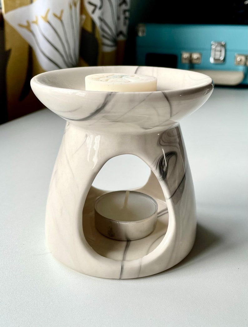 May include: White ceramic wax melt burner with a marble design. The burner has a round top with a small opening in the centre for a tea light. The burner is sitting on a white surface.