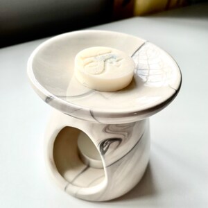 May include: White ceramic wax melt warmer with a marble pattern. The warmer has a round top with a small well for the wax melt and a lower section with a hole for a tea light. A white wax melt is sitting in the well.